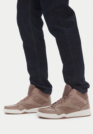 Osoba u svijetlosmeđim tenisicama s visokim vrhom i tamnoplavim trapericama stoji na bijeloj površini.