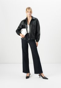 En blond kvinne iført en svart skinnjakke, hvit topp, mørke vide jeans, svarte høye hæler, som holder en hvit håndveske mot en hvit bakgrunn.