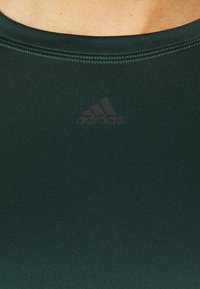 Tummanvihreä urheiluyläosa, jossa on sileä pinta, pyöreä kaula-aukko ja huomaamaton adidas-logo heijastavana painatuksena rinta-alueella.