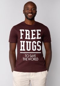 T-shirt bordeaux avec un texte blanc indiquant "CÂLINS GRATUITS POUR SAUVER LE MONDE". Manches courtes, col rond, en matériau coton.