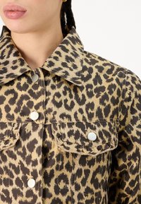 Denim jakna s leopardjim vzorcem, ki vključuje ovratnik, snap gumbke in žepke na prsih; barve vključujejo bež, črno in rjavo.