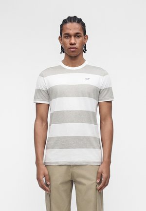 Mann mit geflochtenem Haar, der ein grau-weiß gestreiftes Kurzarmshirt und beige Hosen trägt, steht vor einem schlichten Hintergrund.