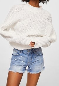 Suéter de punto blanco grueso con cuello alto; combinado con shorts de mezclilla de color azul claro con acentos de perlas y dobladillos deshilachados.
