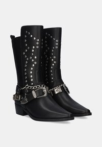 Botas de vaquero de cuero negro con punta afilada, tachuelas plateadas a lo largo del caño y cadenas con hebillas decorativas alrededor del tobillo.