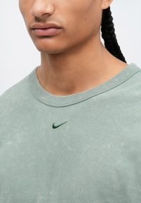 Tricou verde din bumbac, cu guler rotund, prezentând un mic logo Nike brodat în verde închis pe piept. Cânepă moale, texturată.