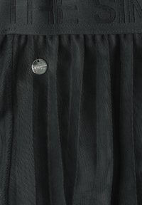 Tissu en maille noire transparente avec texture froncée et un pendentif rond métallique suspendu près de la ceinture élastique supérieure avec texte en relief.