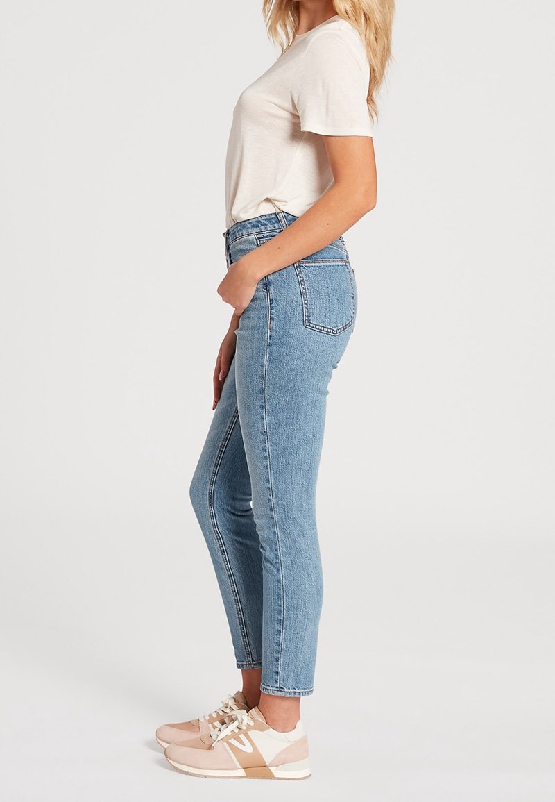 Femme se tenant de côté, portant un t-shirt blanc, un jean slim taille haute bleu clair et des baskets beige-blanc sur un fond uni.