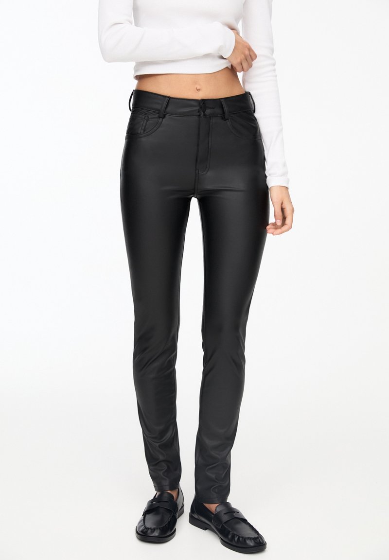 Femme portant un pantalon slim en cuir synthétique noir, un top blanc court à manches longues et des mocassins noirs en cuir, debout devant un fond uni.