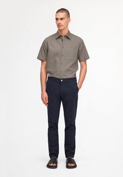 Homme aux cheveux courts portant une chemise grise à manches courtes boutonnée, un pantalon bleu marine et des sandales noires, debout devant un fond blanc.