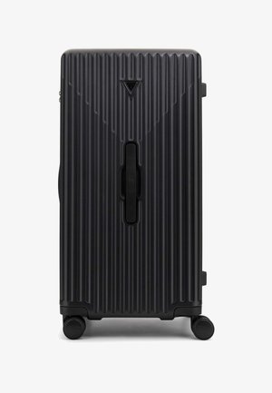 Valise rigide noire avec texture nervurée verticale, une poignée centrale et quatre roues pour la mobilité. Présente un logo en forme de triangle.