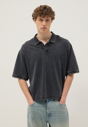 Junger Mann mit lockigem Haar, der ein lässiges dunkelgraues Polohemd und hellblaue Jeans trägt, Hände in den Taschen, neutraler Hintergrund.