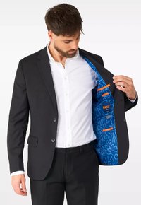 Chaqueta de traje negra con un forro de patrón azul. Cuenta con dos botones, bolsillos laterales y detalles en naranja brillante en el interior. Camisa blanca debajo.