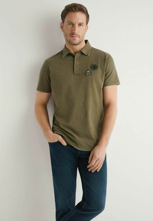 Mężczyzna o krótkich włosach, ubrany w oliwkową koszulkę polo i ciemnoniebieskie dżinsy, stoi przy białej ścianie z jedną ręką w kieszeni.