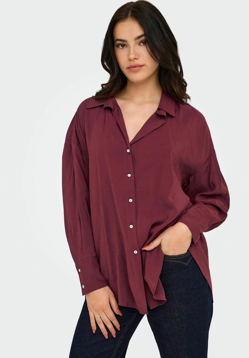Giovane donna con capelli scuri che indossa una camicia button-up bordeaux ampia e jeans scuri, con una mano parzialmente in tasca e un'espressione neutra.