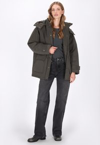 Bergparka in Dunkelgrün mit Reißverschluss, verstellbare Kapuze, großen Fronttaschen und lässiger Passform, kombiniert mit dunklen Jeans.