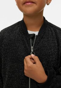 Dziecko trzymające suwak lśniącej czarnej kurtki, ubranej w białą koszulę pod spodem, z widoczną małą złotą kolczykiem w kształcie kółka.