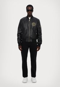 Giubbotto bomber in pelle nera con polsini a costine, chiusura con zip, tasche laterali e patch con logo sul petto. Abbinato a pantaloni neri e sneakers bianche.