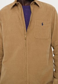 Uomo che indossa una camicia di velluto a coste color beige con zip sopra un maglione viola a trecce con logo ricamato di un giocatore di polo blu sulla tasca del petto.