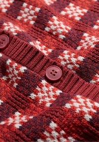 Detailný pohľad na červenú, bielu a tmavofialovú pleteninu s rebrovaným okrajom a dvoma červenými gombíkmi s štyrmi otvormi.