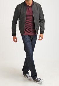 Giacca bomber in pelle grigio scuro, polo bordeaux, jeans blu scuro e sneakers grigie con suola bianca, in uno stile casual mentre cammina.