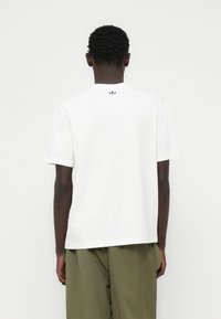 Λευκό βαμβακερό T-shirt με στρογγυλή λαιμόκοψη και κοντά μανίκια· διαθέτει ένα μικρό ναυτικό λογότυπο στο πάνω μέρος της πλάτης. Συνδυάζεται με παντελόνια σε ελιά πράσινο χρώμα.