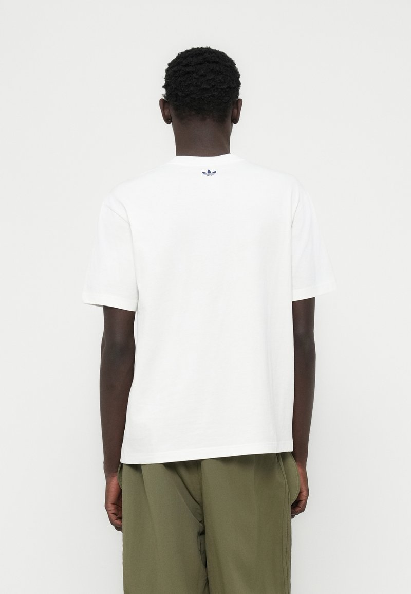 T-shirt en coton blanc avec un col rond et des manches courtes ; présente un petit logo bleu marine sur le haut du dos. Associé à un pantalon vert olive.