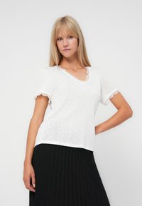 Hvid kortærmet T-shirt med V-hals og blonder på ærmerne og ved halsudskæringen. Stoffet virker let og har en let tekstur.