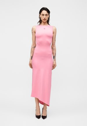 Persona con tatuaggi indossa un vestito midi rosa senza maniche e tacchi neri, in piedi davanti a uno sfondo grigio chiaro uniforme.