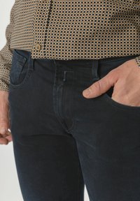 Mørkeblå denim jeans med en glatt tekstur, som har en frontlomme og en slank passform. En rutete skjorte i brunt og hvitt er delvis synlig ovenfor.