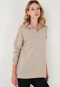 Beige színű cipzáras pulóver, magas gallérral, hosszú ujjakkal és oldalsó hasadékokkal. Simás textúrájú és laza szabású. Fekete leggings van rajta.