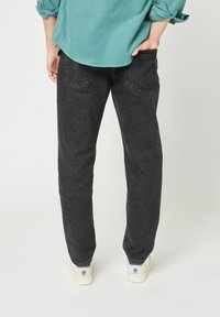 Persona che indossa jeans neri, camicia a maniche lunghe turchese, sneakers bianche, in piedi con una mano nella tasca posteriore contro uno sfondo chiaro uniforme.