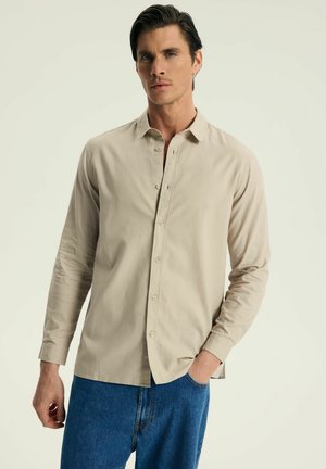 Beżowa koszula z długim rękawem, z kołnierzykiem typu button-down, z przodu zapinana na guziki, o luźnym kroju, zestawiona z niebieskimi dżinsami. Gładka textura materiału.