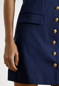 Vestido azul-marinho com tecido texturizado, um bolso de aplicação e quatro botões dourados decorativos na frente para fecho.