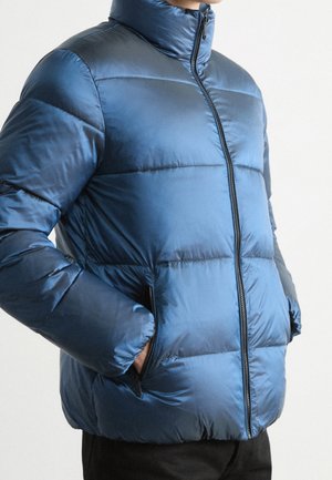Persona che indossa una giacca imbottita blu lucida con colletto alto, cerniera frontale e mani nelle tasche laterali con cerniera.