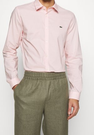 Mężczyzna w jasnoróżowej koszuli na guziki z długimi rękawami i oliwkowych spodniach z elastycznym pasem, stojący z lewą ręką w kieszeni.