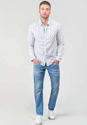 Camisa blanca de botones con pequeño patrón floral azul, combinada con jeans azul claro y zapatillas blancas. Diseño simple y casual.