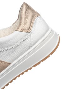 Bílá kožená teniska s béžovým kovovým detailem na patě, texturovanou bílou podrážkou a béžovou gumovou podešví, zobrazená v detailním záběru.