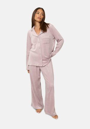Femme portant un haut de pyjama en velours rose pâle à boutons et manches longues, avec un pantalon large assorti, debout pieds nus sur un fond blanc.