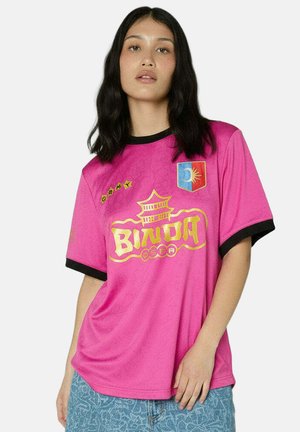 Femme aux longs cheveux noirs portant un maillot de sport rose avec des lettres jaunes et un jean bleu sur un fond blanc.