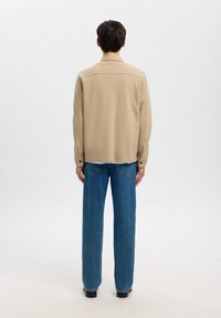 Homme debout, dos tourné, portant une veste beige, un jean bleu et des chaussures noires sur un fond clair uni.
