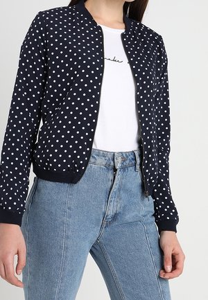 Femme portant une veste zippée bleu marine à pois blancs par-dessus une chemise blanche et un jean taille haute bleu clair.