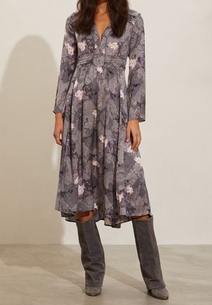 Femme portant une robe midi grise à fleurs avec manches longues et col en V, assortie à de hautes bottes en daim gris sur un fond uni.