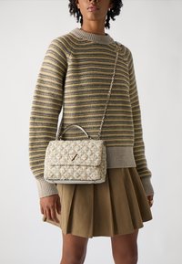 Borsa a tracolla trapuntata color crema e oro con forma strutturata e manico superiore. Presenta un logo triangolare in tonalità oro e una tracolla a catena, abbinata a un maglione a righe.