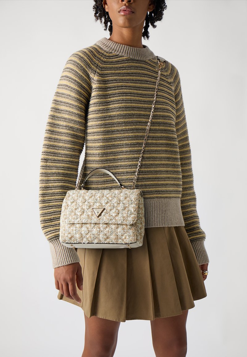 Borsa a tracolla trapuntata color crema e oro con forma strutturata e manico superiore. Presenta un logo triangolare in tonalità oro e una tracolla a catena, abbinata a un maglione a righe.
