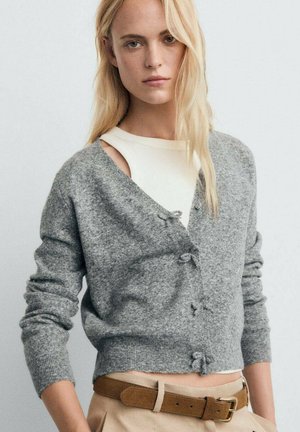 Cardigan gris en tricot avec un col en V, des boutons sur le devant et des poignets côtelés. Porté sur un haut blanc avec une texture distincte. Coupe décontractée.