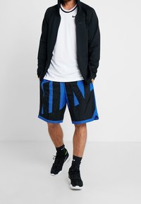 Sudadera con cremallera negra, camiseta deportiva blanca, pantalones cortos azules y negros con gráficos en negrita de "NIKE", combinados con zapatillas negras y calcetines negros.