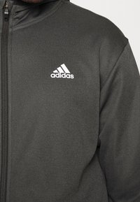 Sudadera con cremallera negra de tejido texturizado, con un logo blanco de Adidas en el área del pecho izquierdo y una capucha.
