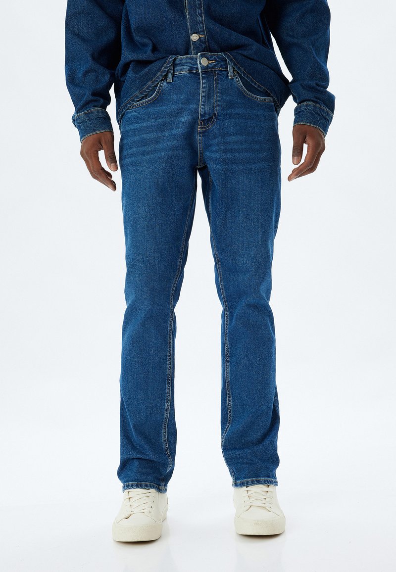 Jeans de mezclilla azules con corte recto, con costuras en contraste, cierre de botón y bolsillos frontales. Llevados con zapatillas blancas.