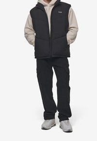 Czarna kamizelka puchowa z zamkiem, noszona na jasnoszarej bluzie z kapturem. Uzupełniona czarnymi spodniami cargo i jasnymi butami, które mają subtelne logo.