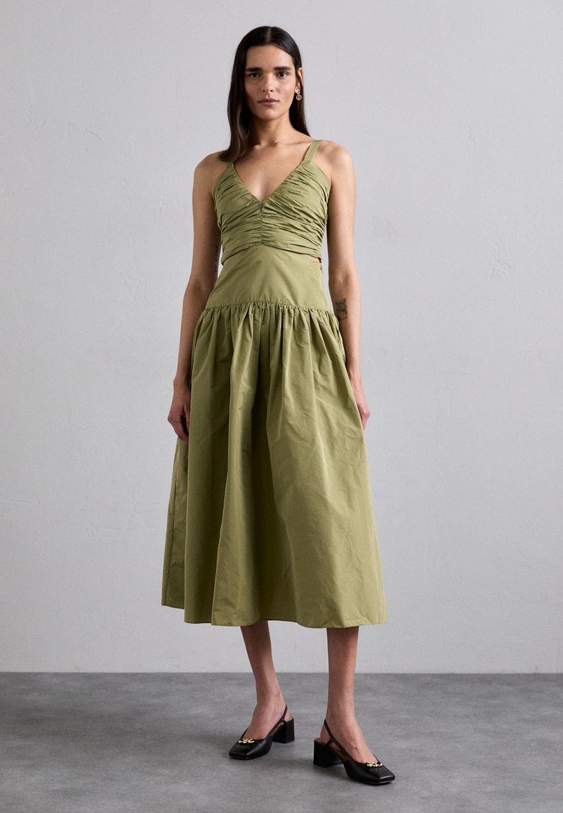 Vestido verde oliva con un cuerpo fruncido y una falda acampanada y recogida. Combinado con zapatos negros de tacón cuadrado. Textura suave, diseño minimalista.